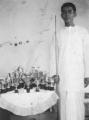 Photo of Chandra Hirjee, dressed in bush shirt and dhoti, with his early collection of trophies